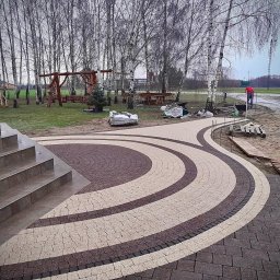 Rosa Ogrody - Okrągła ścieżka z kostki brukowej w dwóch kolorach, prowadząca od schodów w kierunku ogrodu z drzewami i elementami placu zabaw. W tle osoba w czerwonej koszulce.