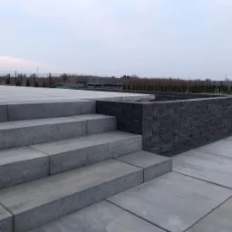Nowoczesne schody i mur z ciemnej kostki, wykonane z precyzją. Szare płyty chodnikowe tworzą minimalistyczny design w przestrzeni zewnętrznej. Widok na ogród w tle.