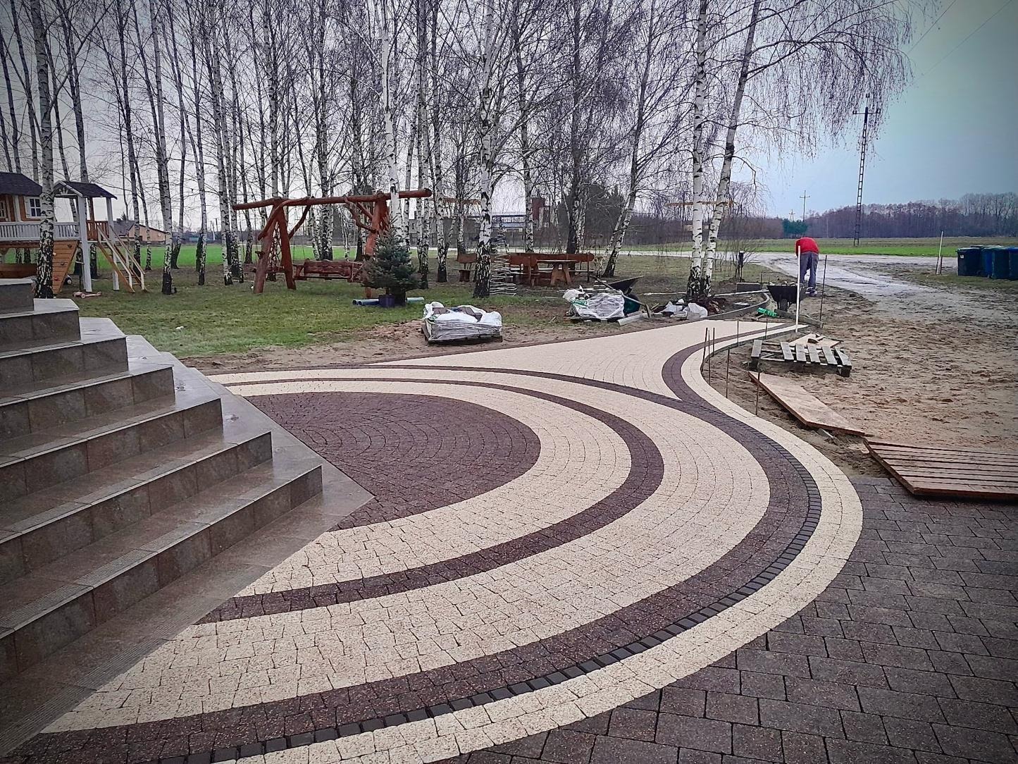Okrągła ścieżka z kostki brukowej w dwóch kolorach, prowadząca od schodów w kierunku ogrodu z drzewami i elementami placu zabaw. W tle osoba w czerwonej koszulce.