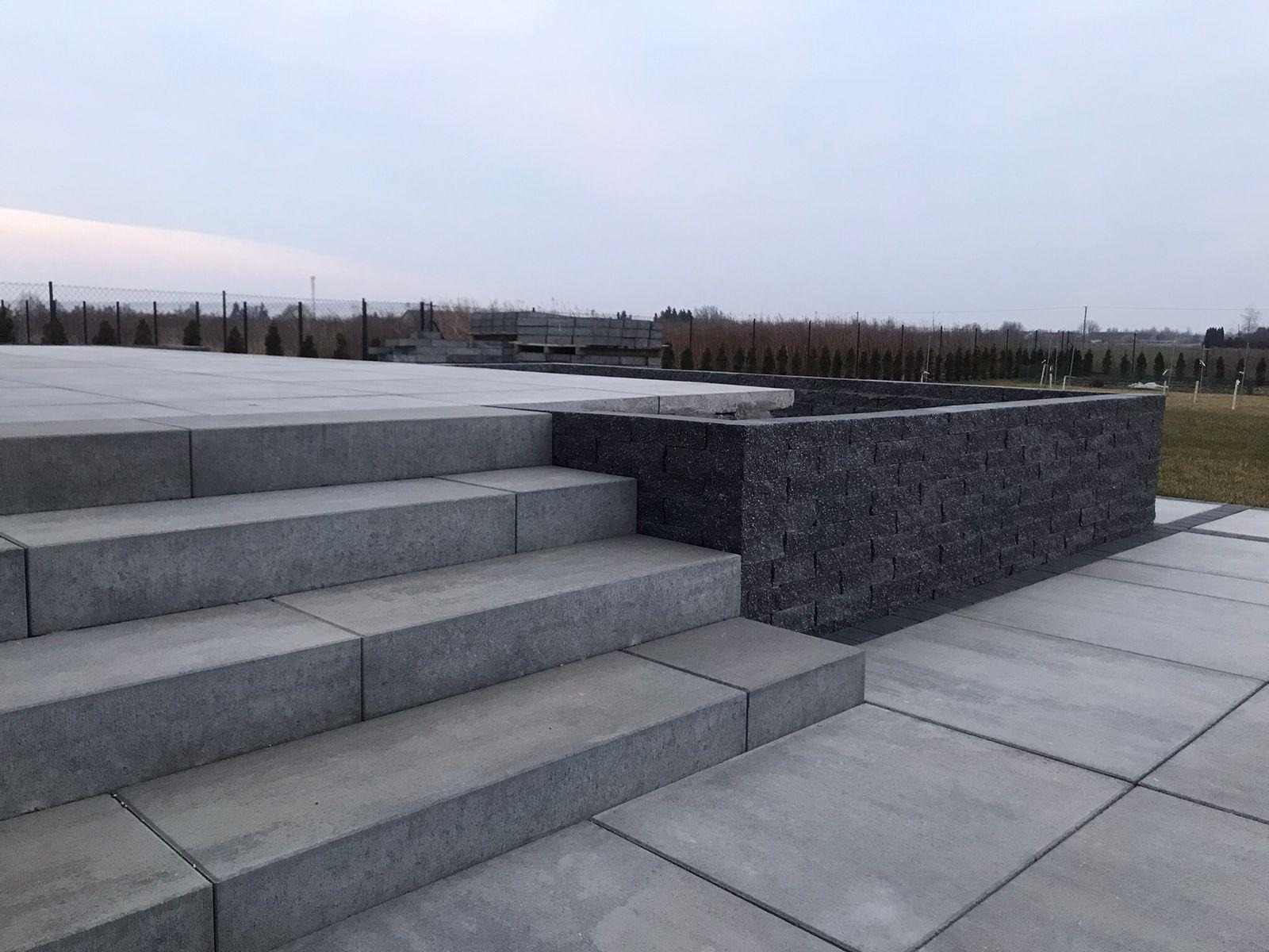 Nowoczesne schody i mur z ciemnej kostki, wykonane z precyzją. Szare płyty chodnikowe tworzą minimalistyczny design w przestrzeni zewnętrznej. Widok na ogród w tle.