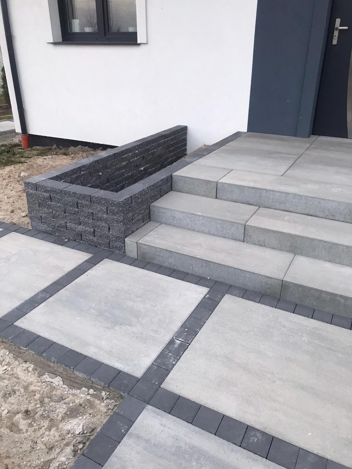 Nowoczesne schody wejściowe i ścieżka z jasnoszarej kostki brukowej, obrzeża z ciemnej kostki. Donica z ciemnej cegły przy wejściu do domu.