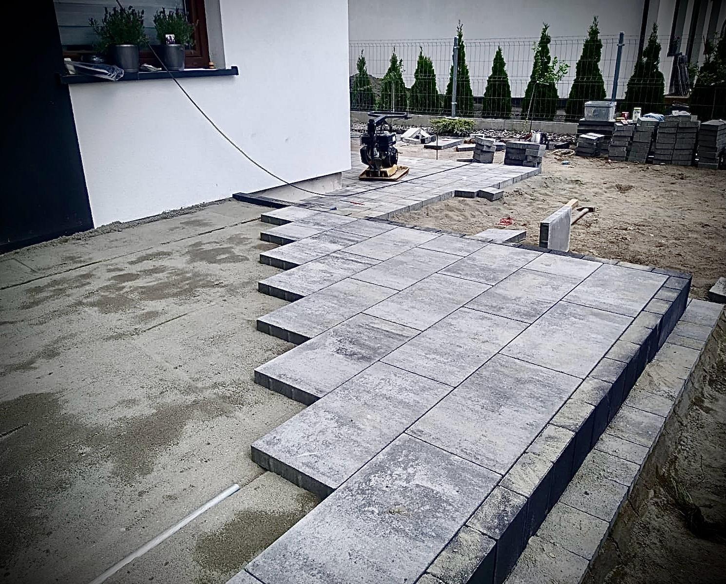 Układanie szarej kostki brukowej na posesji domu jednorodzinnego. Widoczna ubijarka, zapas kostki i przygotowane podłoże. Częściowo ułożony chodnik z kostki.