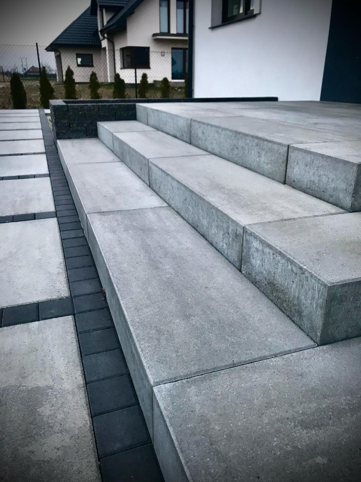 Schody i chodnik z szarej kostki brukowej, obrzeża z ciemnej kostki. Nowoczesny design, precyzyjne ułożenie, dom w tle. Minimalistyczna estetyka.