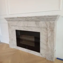 Marmurowa obudowa kominka elektrycznego w nowocześnie wykończonym salonie z parkietem w jodełkę i sztukaterią na ścianach.