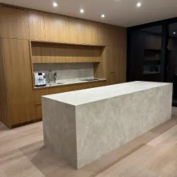 Nowoczesna kuchnia z wyspą wykończoną beżowym materiałem imitującym marmur, drewniane szafki w tle i parkiet na podłodze.