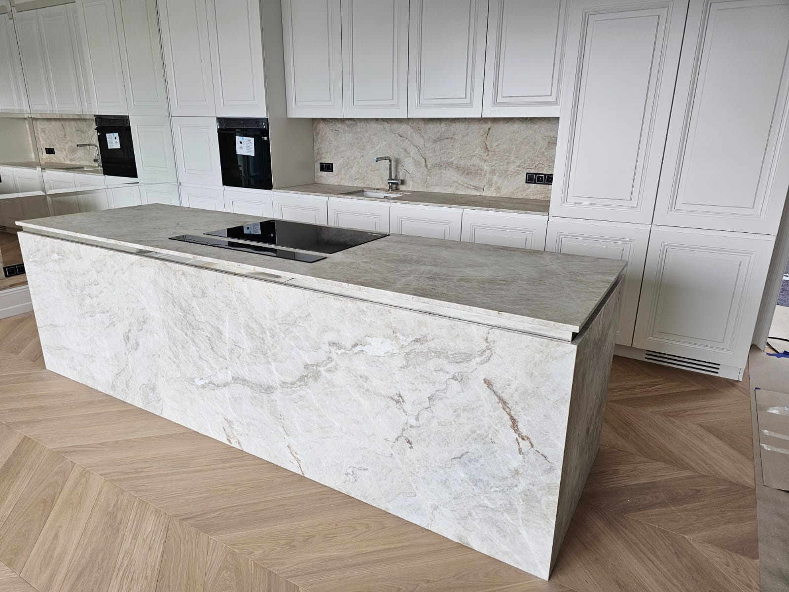 Wyspa kuchenna z blatem z kamienia naturalnego o jasnym, marmurkowym wzorze, z wbudowaną płytą indukcyjną, na tle białych szafek kuchennych i parkietu ułożonego w jodełkę.