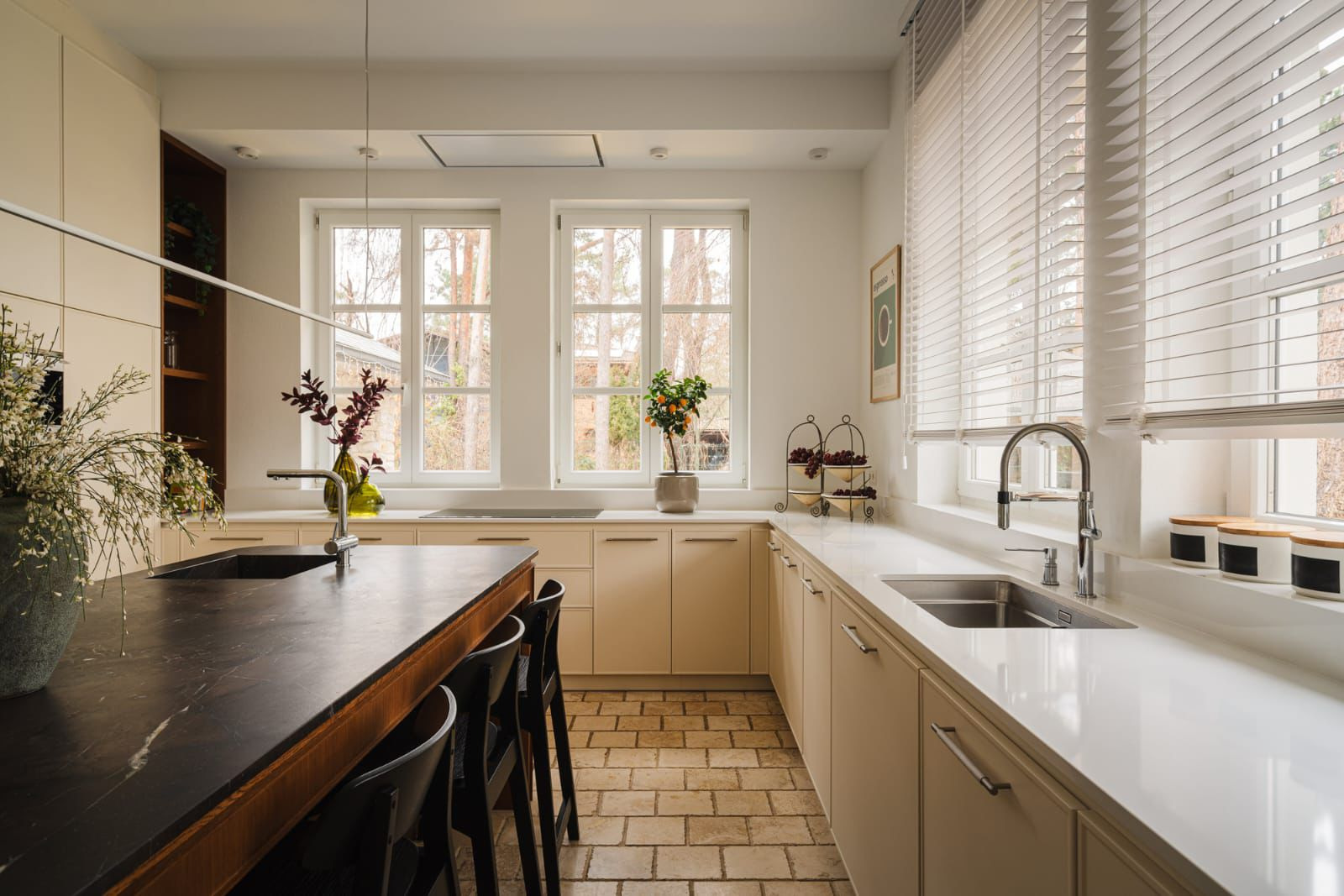 Jasna kuchnia z ciemnym kamiennym blatem wyspy, ceglaną podłogą, beżowymi szafkami i białym blatem pod oknem z żaluzjami.