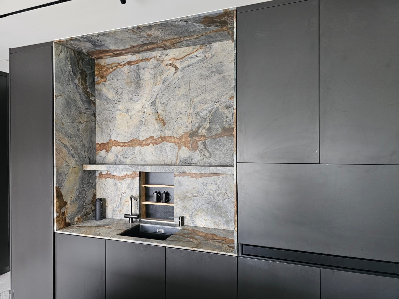 Nowoczesna kuchnia z blatem z kamienia naturalnego o wyraźnym wzorze, dopasowanym do obudowy wnęki z półką, czarny zlew i matowe, grafitowe szafki.