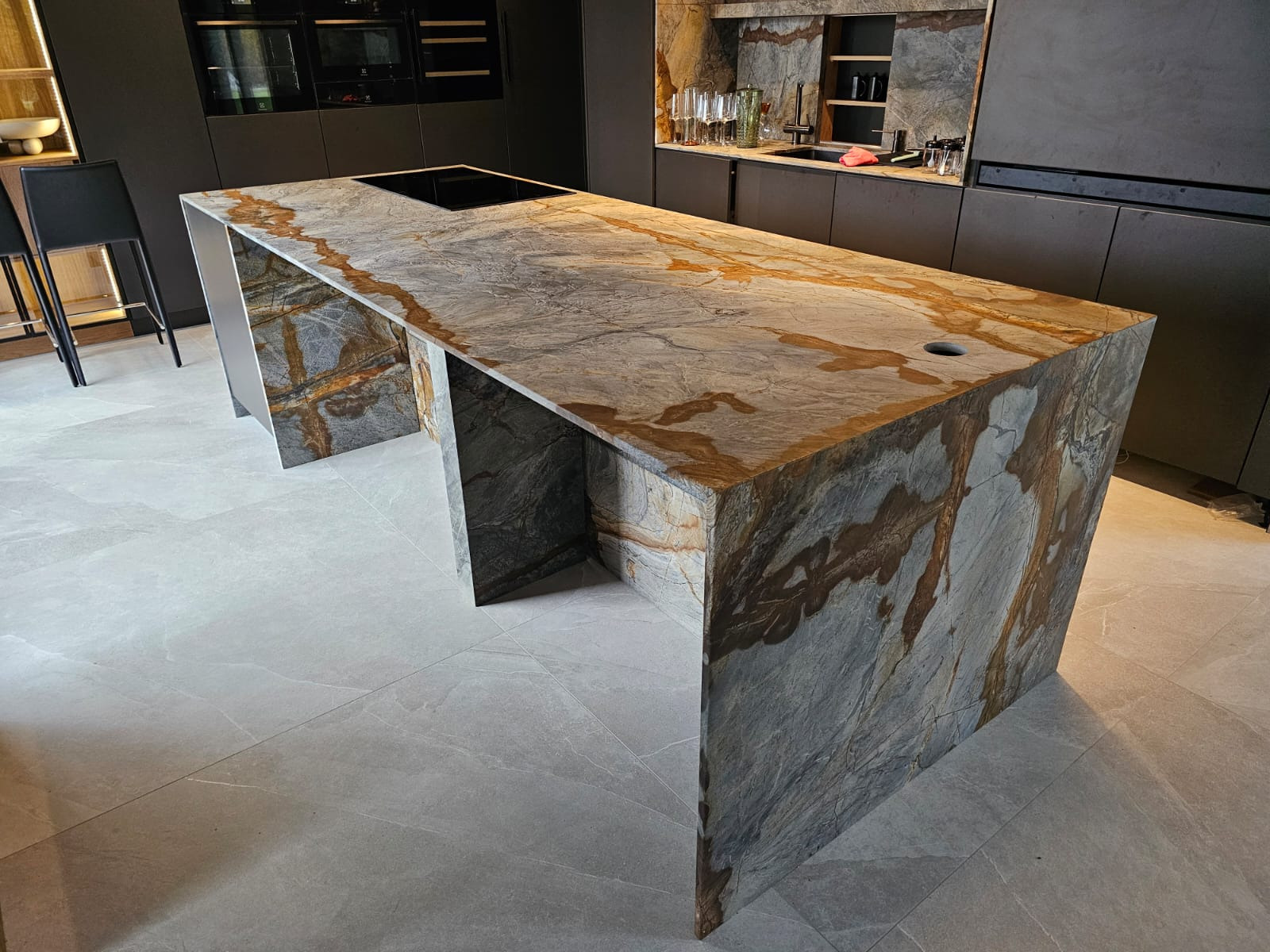 Kuchenna wyspa z blatem z kamienia naturalnego o wyraźnym, warstwowym wzorze w odcieniach szarości i brązu, z wbudowaną płytą grzewczą i otworem na zlew, na tle nowoczesnej zabudowy kuchennej...