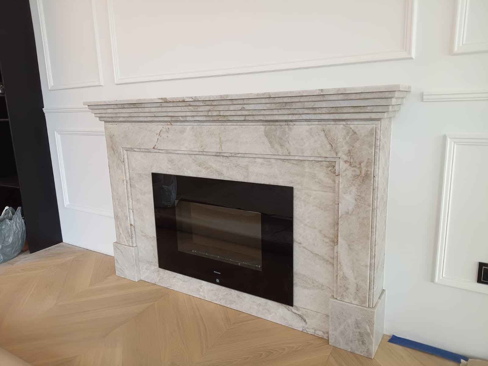 Marmurowa obudowa kominka elektrycznego w nowocześnie wykończonym salonie z parkietem w jodełkę i sztukaterią na ścianach.