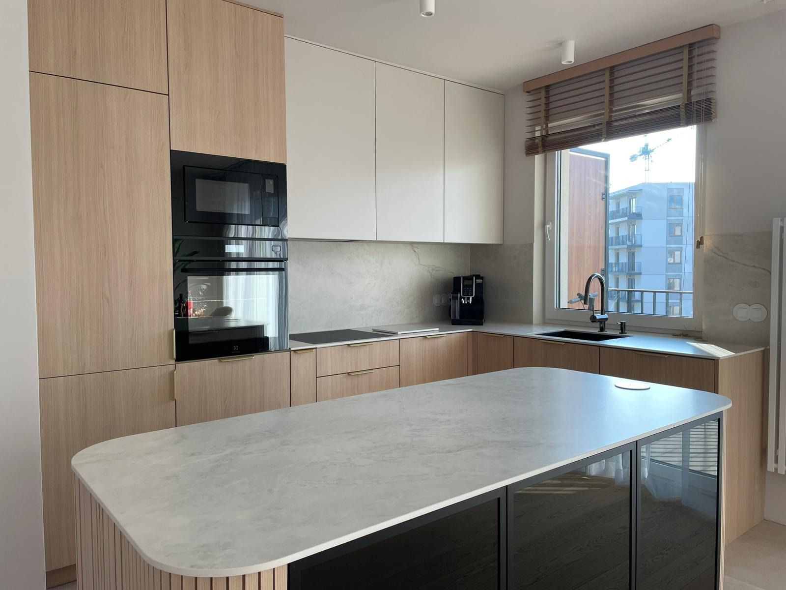 Jasna kuchnia z wyspą i blatem z szarego kamienia, fronty szafek z jasnego drewna, zabudowa kuchenna pod sufit, okno z drewnianymi żaluzjami.