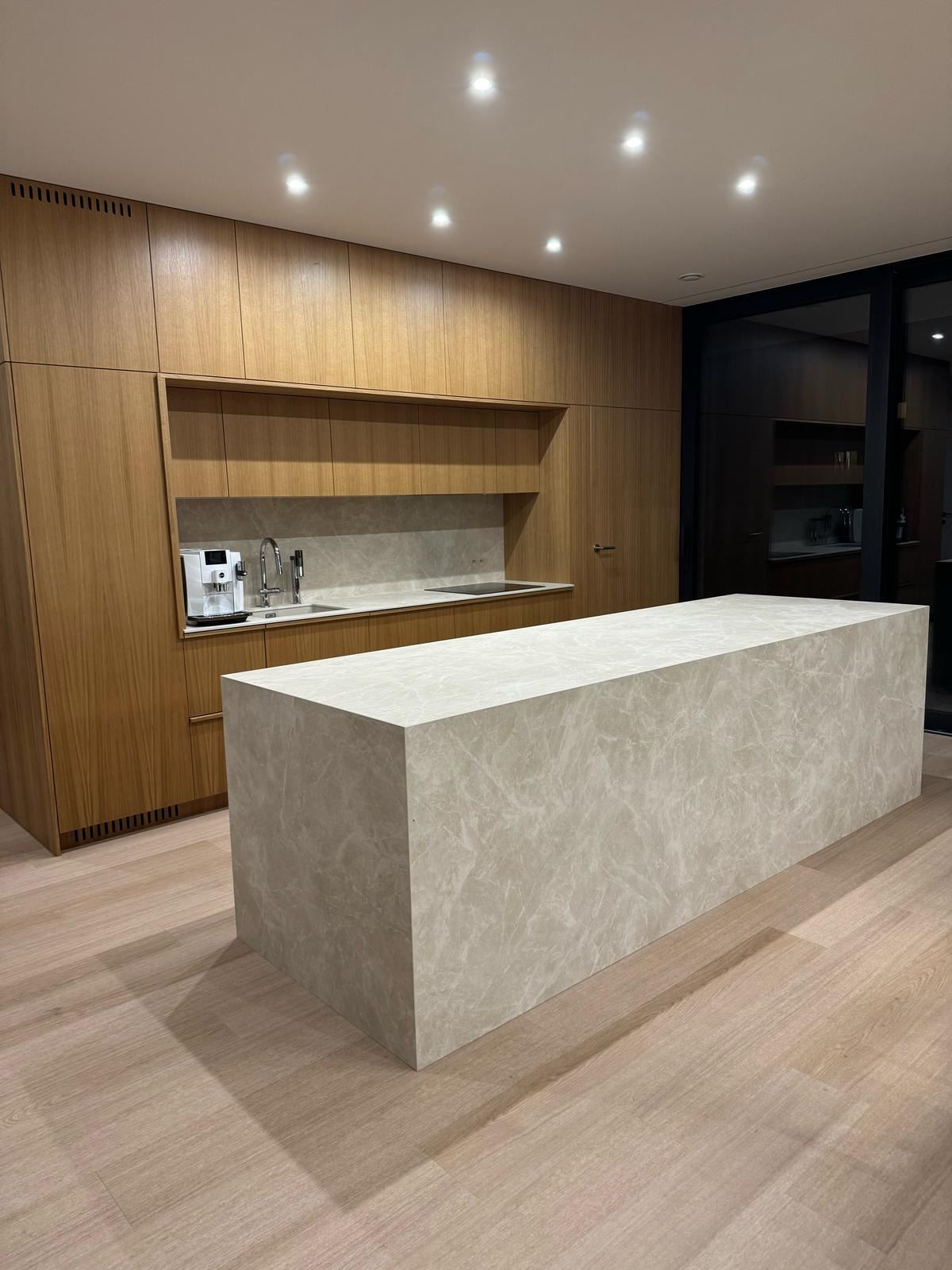 Nowoczesna kuchnia z wyspą wykończoną beżowym materiałem imitującym marmur, drewniane szafki w tle i parkiet na podłodze.