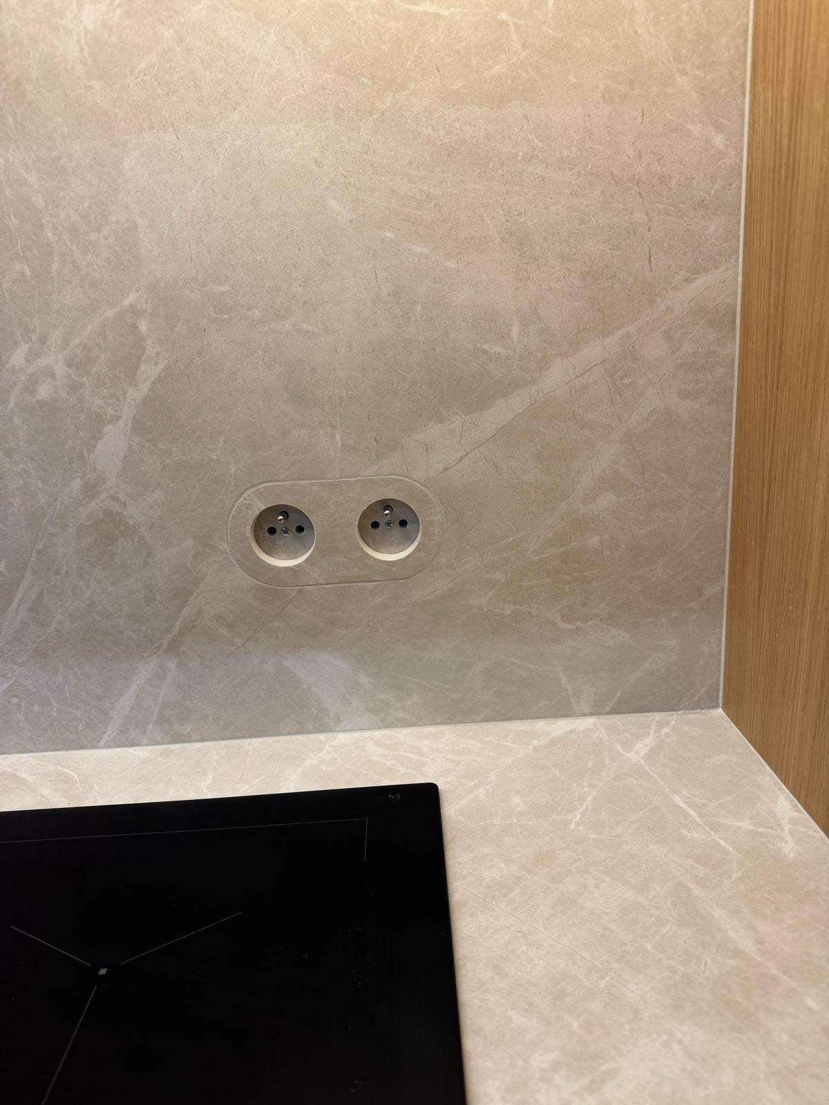 Beżowy blat kamienny z marmurkowym wzorem i dopasowaną okładziną ścienną, w której zamontowano podwójne gniazdko elektryczne nad czarną płytą indukcyjną.