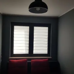 Pokój z ciemnoszarymi ścianami, oknem z żaluzjami poziomymi i dwoma czerwonymi poduszkami na kanapie, oświetlony lampą sufitową.