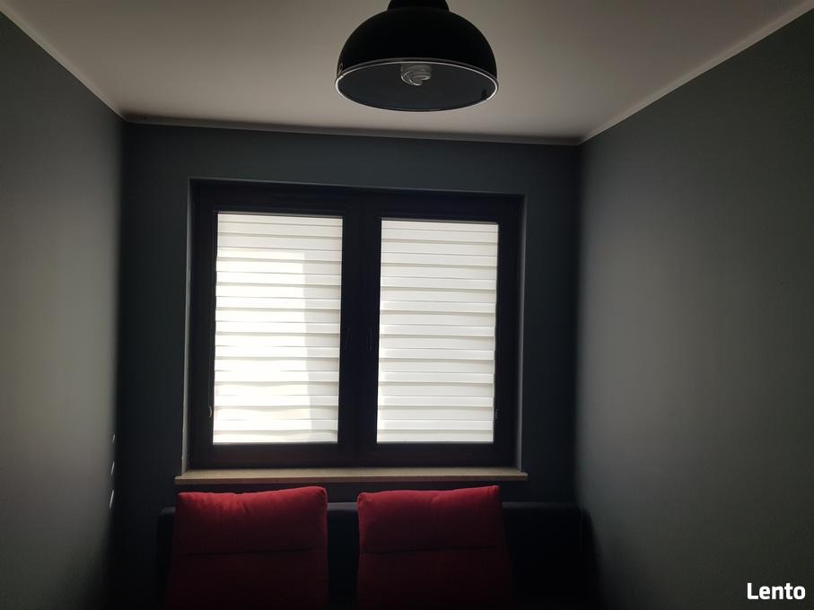 Pokój z ciemnoszarymi ścianami, oknem z żaluzjami poziomymi i dwoma czerwonymi poduszkami na kanapie, oświetlony lampą sufitową.