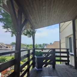 Zadaszony taras drewniany z ciemnymi belkami i deskami, widok na ogród i domy, wisząca donica z białymi kwiatami, donica z zielonymi roślinami w rogu.