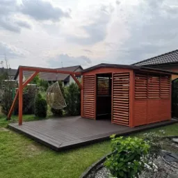 Zadaszona altana z drewnianymi lamelami, taras kompozytowy w kolorze grafitowym i wiszące krzesło na drewnianej konstrukcji w ogrodzie.