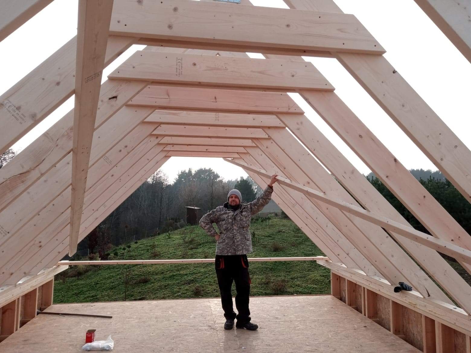 Konstrukcja drewniana dachu altany w trakcie budowy, widoczny pracownik w ubraniu roboczym na tle zielonego wzgórza.