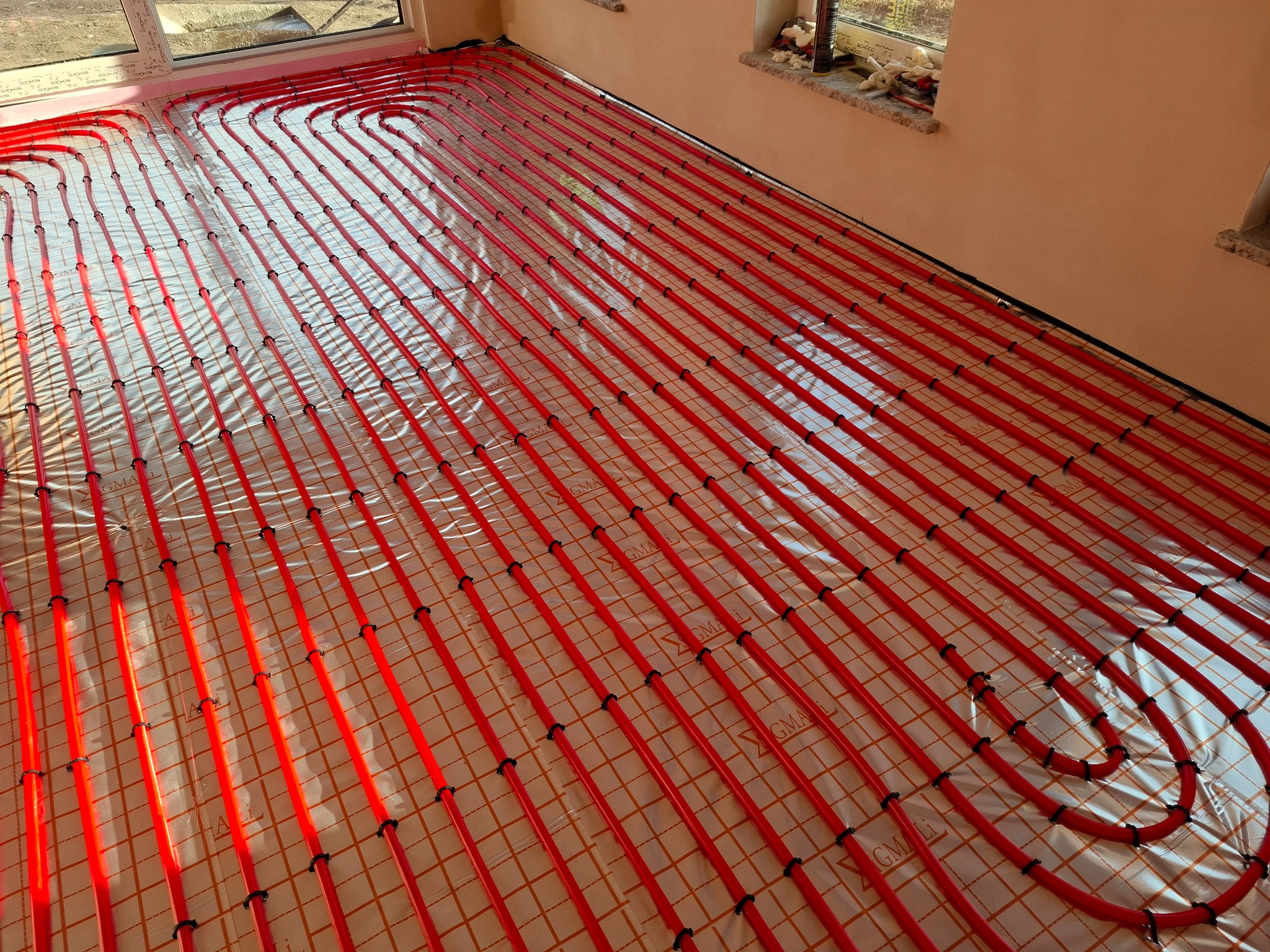 Czerwone rury ogrzewania podłogowego ułożone na folii z siatką, widoczne czarne uchwyty mocujące, jasne wnętrze w trakcie prac instalacyjnych.