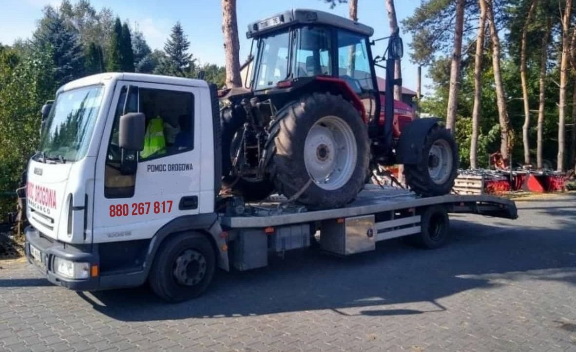 Biała laweta z napisem 'Pomoc Drogowa' przewozi czerwony traktor na tle drzew.