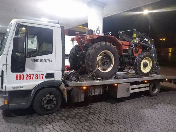 Czerwony traktor z ładowaczem na platformie białej lawety pomocy drogowej z widocznym numerem telefonu, sfotografowany w nocy przy sztucznym oświetleniu.