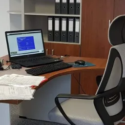 Biuro z biurkiem, na którym stoi laptop z widocznym dokumentem, kalkulator, przybory biurowe i sterta dokumentów. W tle regał z segregatorami i drukarka.