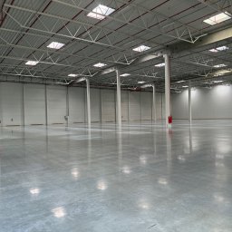 CleanQ - Rozległa, pusta hala przemysłowa z polerowaną, szarą posadzką żywiczną, wsparta na białych kolumnach z czerwonymi elementami. Górne okna wpuszczają naturalne światło.