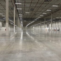 CleanQ - Rozległa, pusta hala magazynowa z betonową posadzką, regularnym układem kolumn i rzędami świetlówek pod sufitem. Widoczne bramy przemysłowe i instalacje przeciwpożarowe.