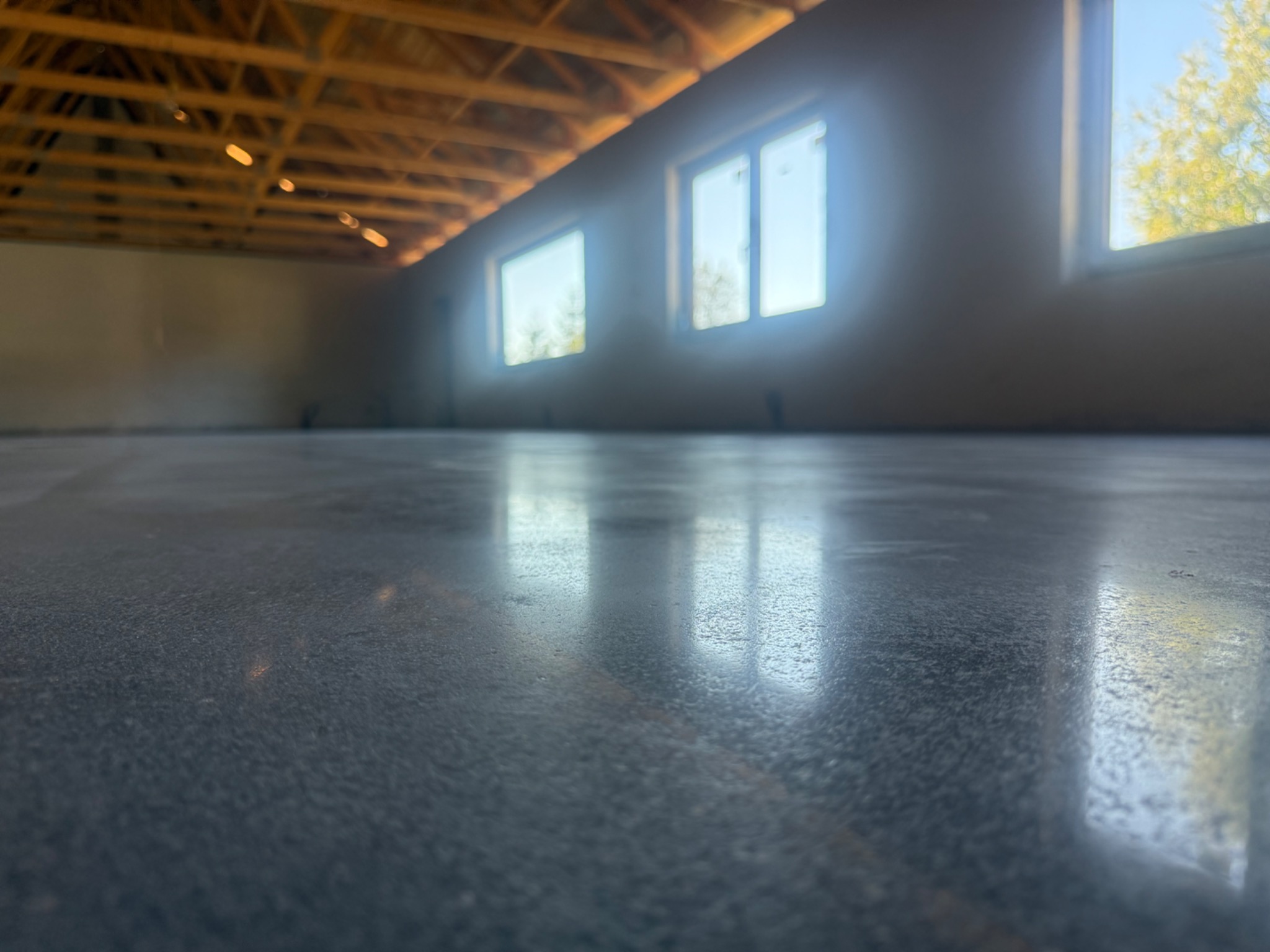 Błyszcząca, szara posadzka przemysłowa z widocznym odbiciem okien i drewnianą konstrukcją stropu w surowym wnętrzu, oświetlonym naturalnym światłem.
