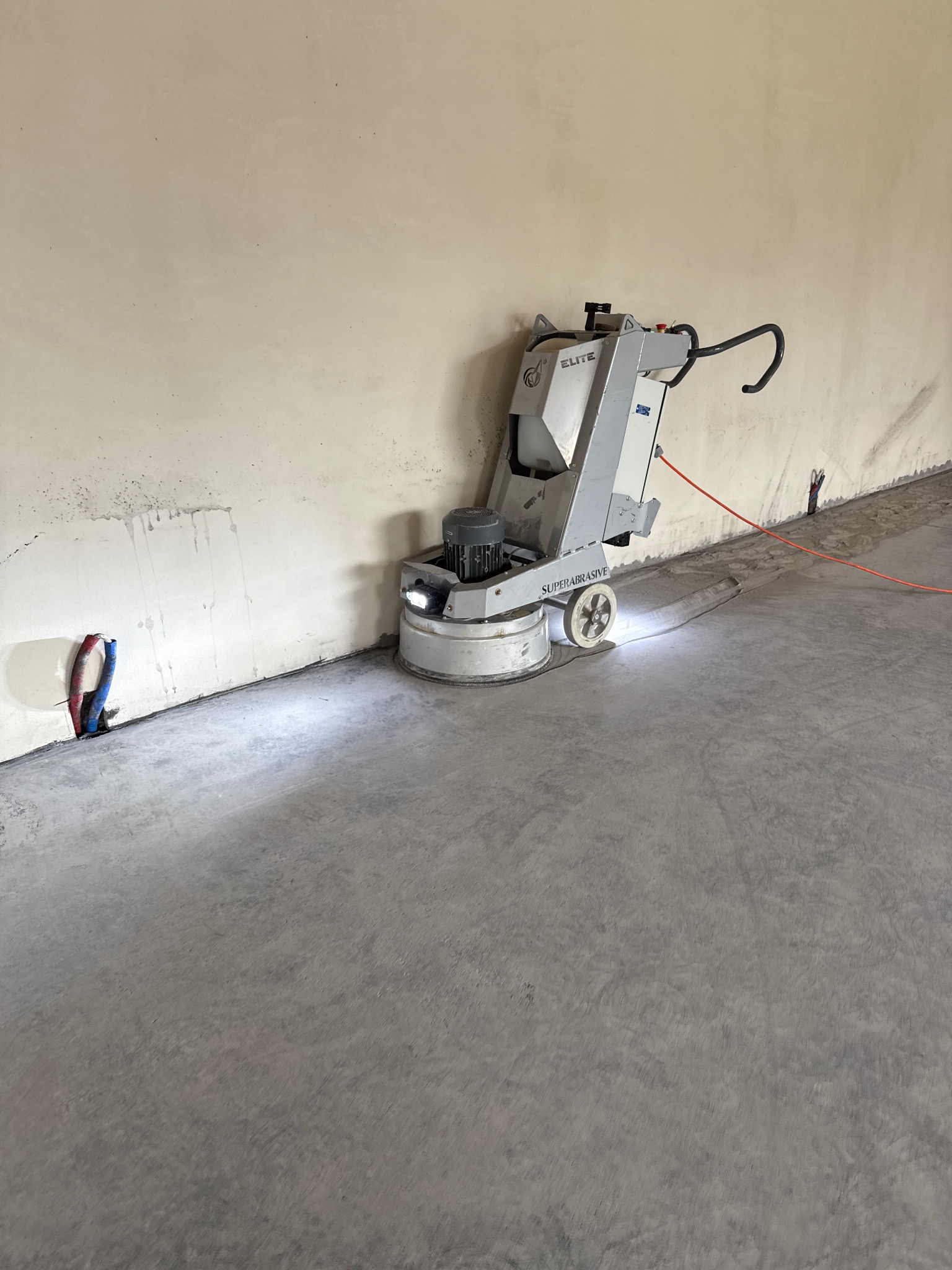 Szlifowanie betonowej posadzki za pomocą szlifierki planetarnej w surowym wnętrzu. Widoczne ślady po szlifowaniu. W tle ściana z instalacjami.