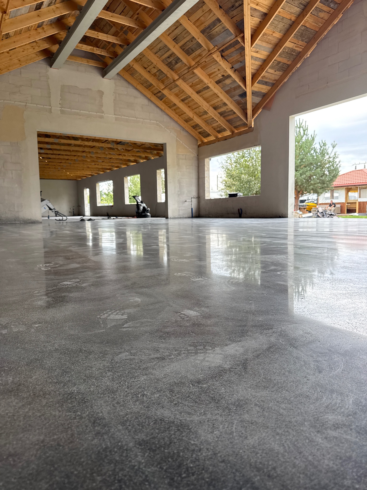 Błyszcząca, szara posadzka w surowym wnętrzu budynku w trakcie budowy. Widoczne ślady, maszyny do obróbki betonu i drewniany strop dachu.