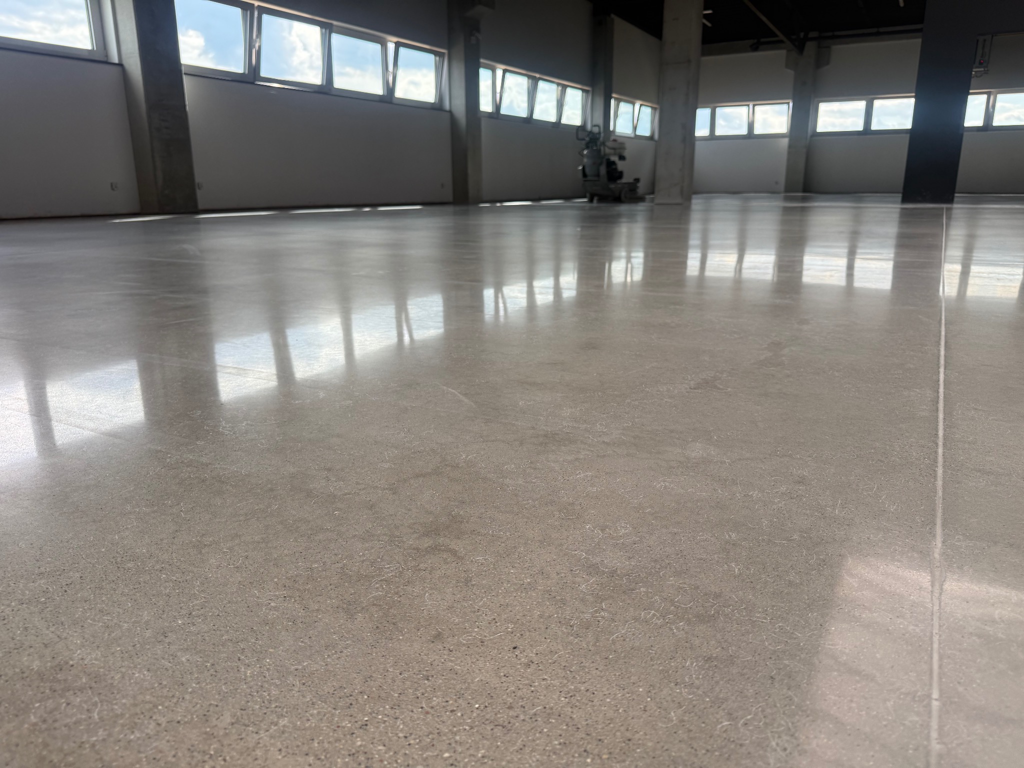Błyszcząca, polerowana posadzka przemysłowa w przestronnym wnętrzu z oknami. Widoczna maszyna do polerowania w oddali. Minimalistyczny design i betonowe filary.