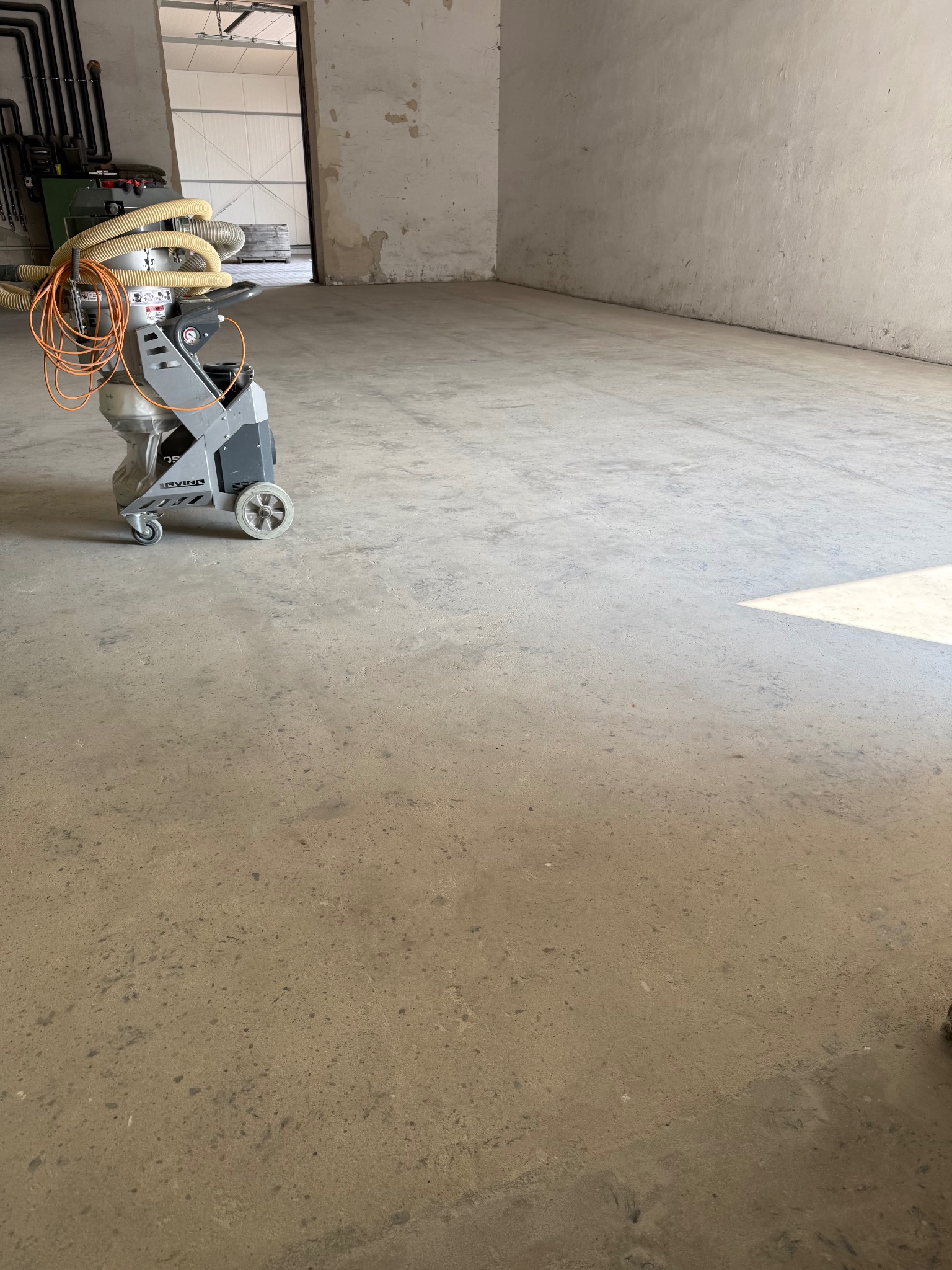 Maszyna do czyszczenia posadzek przemysłowych na betonowej podłodze w jasnym, pustym pomieszczeniu. Widoczne rury i kable maszyny. Oświetlenie dzienne.