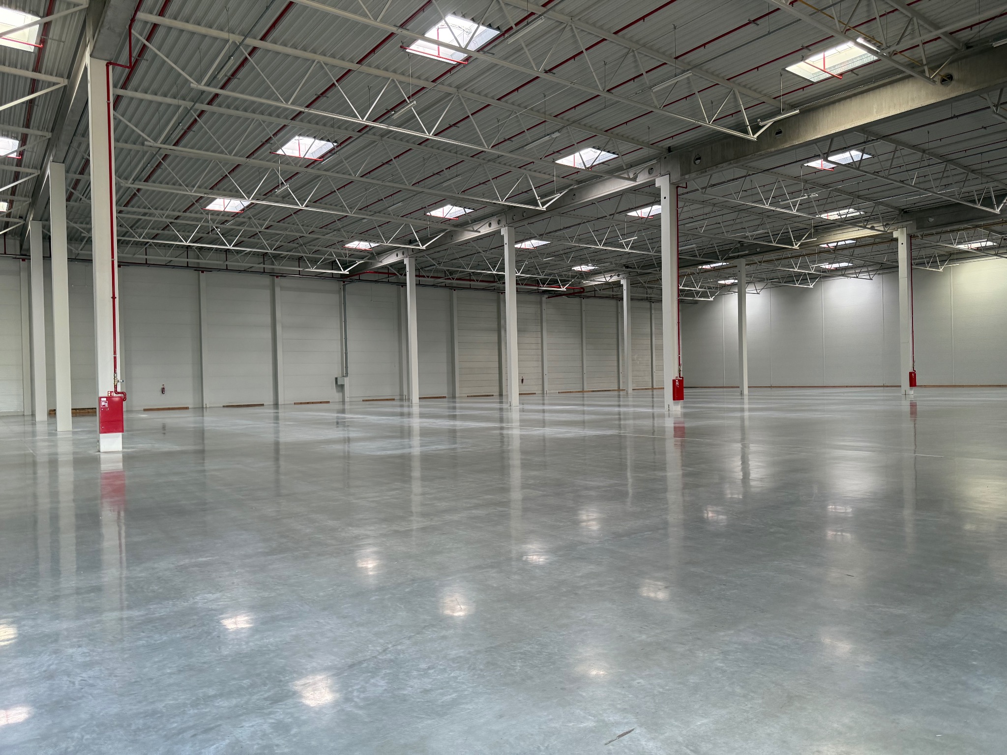 Rozległa, pusta hala przemysłowa z polerowaną, szarą posadzką żywiczną, wsparta na białych kolumnach z czerwonymi elementami. Górne okna wpuszczają naturalne światło.