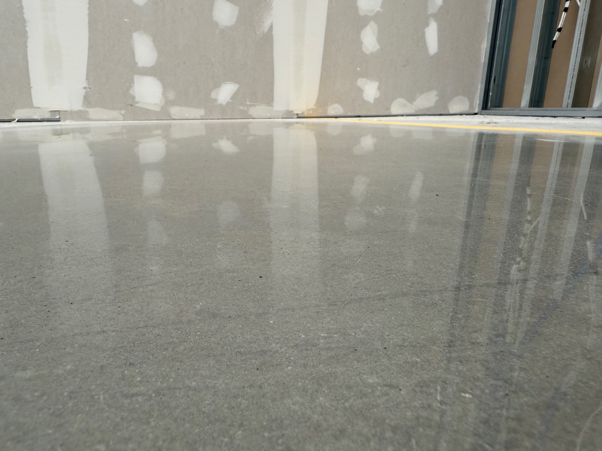 Błyszcząca, szara posadzka przemysłowa z widoczną linią dylatacyjną i fragmentem ściany w trakcie wykańczania, ukazująca gładką powierzchnię i minimalistyczny design.