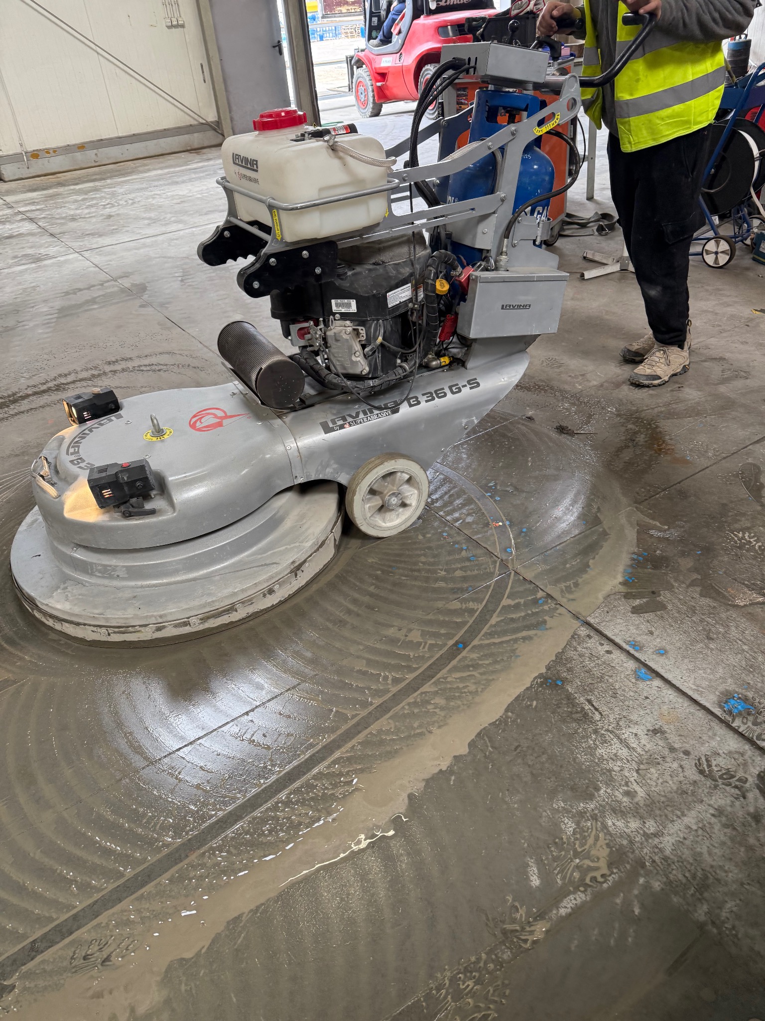 Mycie posadzki przemysłowej maszyną Levina B36 G-S. Widoczne ślady po czyszczeniu, operator w odblaskowej kamizelce. W tle wózek widłowy.