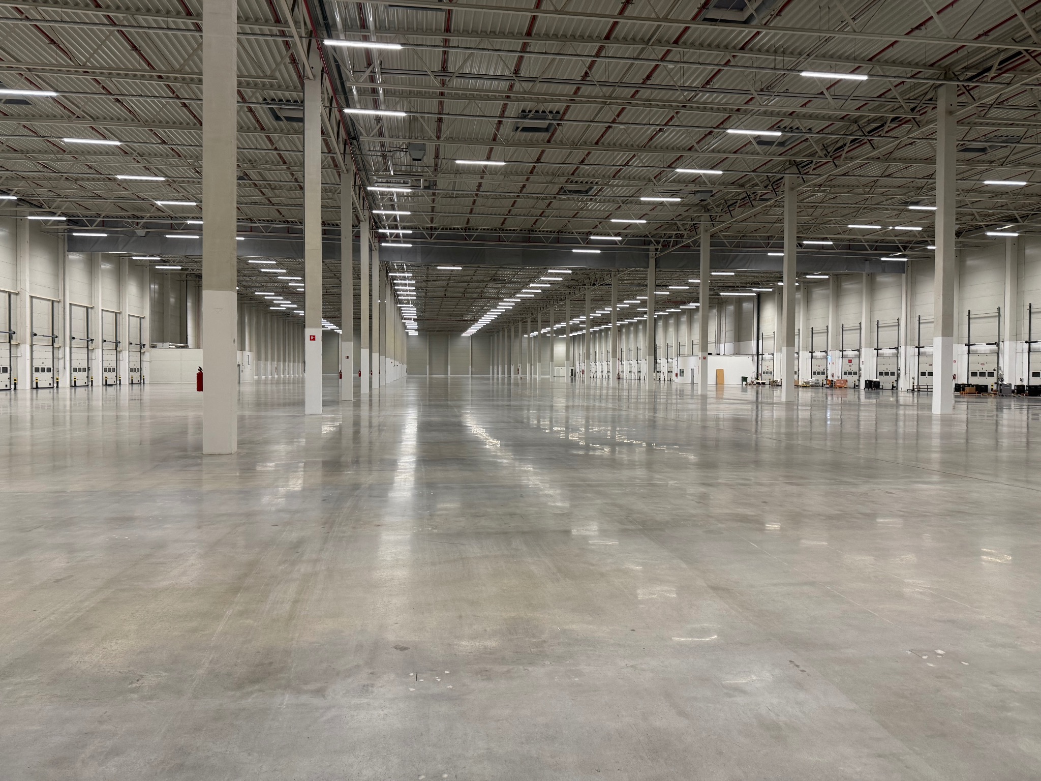 Rozległa, pusta hala magazynowa z betonową posadzką, regularnym układem kolumn i rzędami świetlówek pod sufitem. Widoczne bramy przemysłowe i instalacje przeciwpożarowe.