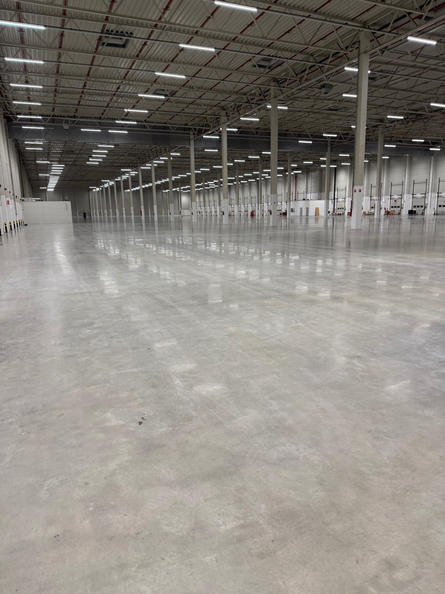 Lśniąca, szara posadzka przemysłowa w przestronnej hali magazynowej z rzędami kolumn i oświetleniem sufitowym. Widok na czystą, gładką powierzchnię.