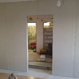 Montaż płyt karton gips na ścianie z widocznym otworem drzwiowym i elementami instalacji elektrycznej w tle.