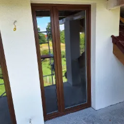 Nowe okna balkonowe z brązowymi ramami w świeżo otynkowanej, białej elewacji budynku w trakcie wykończenia, widoczny fragment dachu z brązową dach&oacute;wką