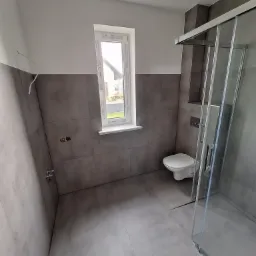 Nowoczesna łazienka z szarymi płytkami, kabiną prysznicową ze szkła i przesuwnymi drzwiami, wiszącą toaletą i oknem z widokiem na dom.