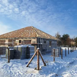 Budowa domu jednorodzinnego w zimowej scenerii: surowy stan, wzniesione mury z bloczków betonowych, drewniana konstrukcja dachu, rusztowania i zapasy materiałów budowlanych przykryte śniegiem...