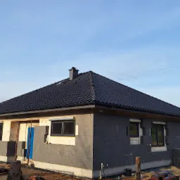 Dom jednorodzinny w trakcie ocieplania, widoczna grafitowa izolacja ścian zewnętrznych i czarna dachówka ceramiczna na skośnym dachu.