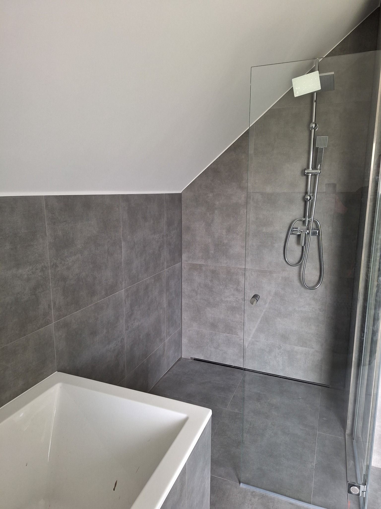 Nowoczesna łazienka na poddaszu z szarymi płytkami, kabiną prysznicową typu walk-in i białą wanną narożną.