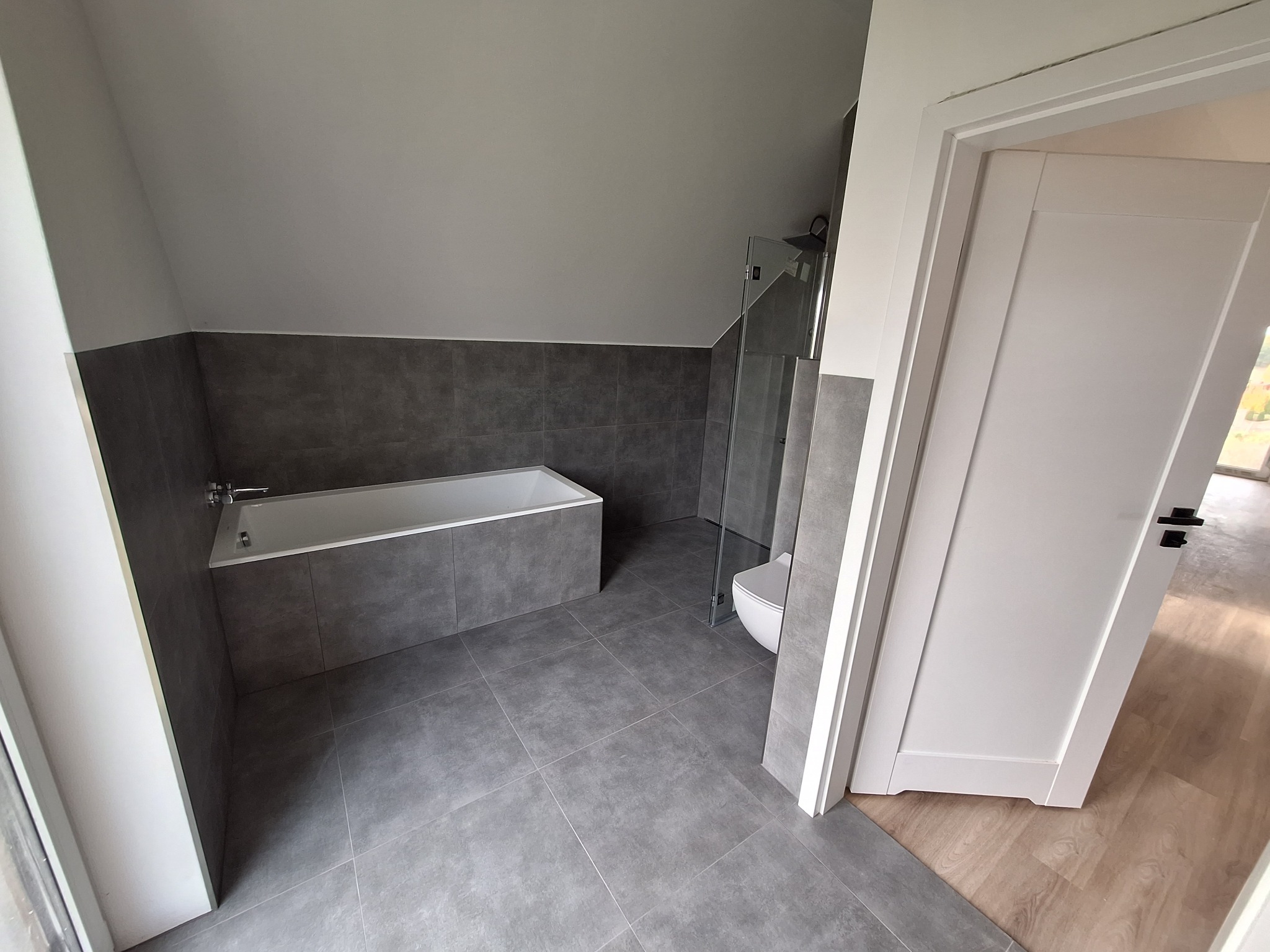 Nowoczesna łazienka na poddaszu z szarą glazurą, białą wanną i kabiną prysznicową ze szklaną ścianką, widok zza uchylonych białych drzwi.