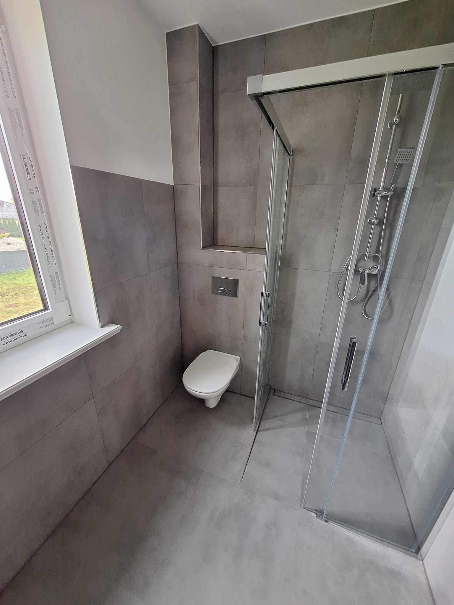 Nowoczesna łazienka z szarymi płytkami, kabiną prysznicową z przezroczystego szkła i podwieszaną toaletą, widziana z perspektywy narożnej, z oknem w tle.