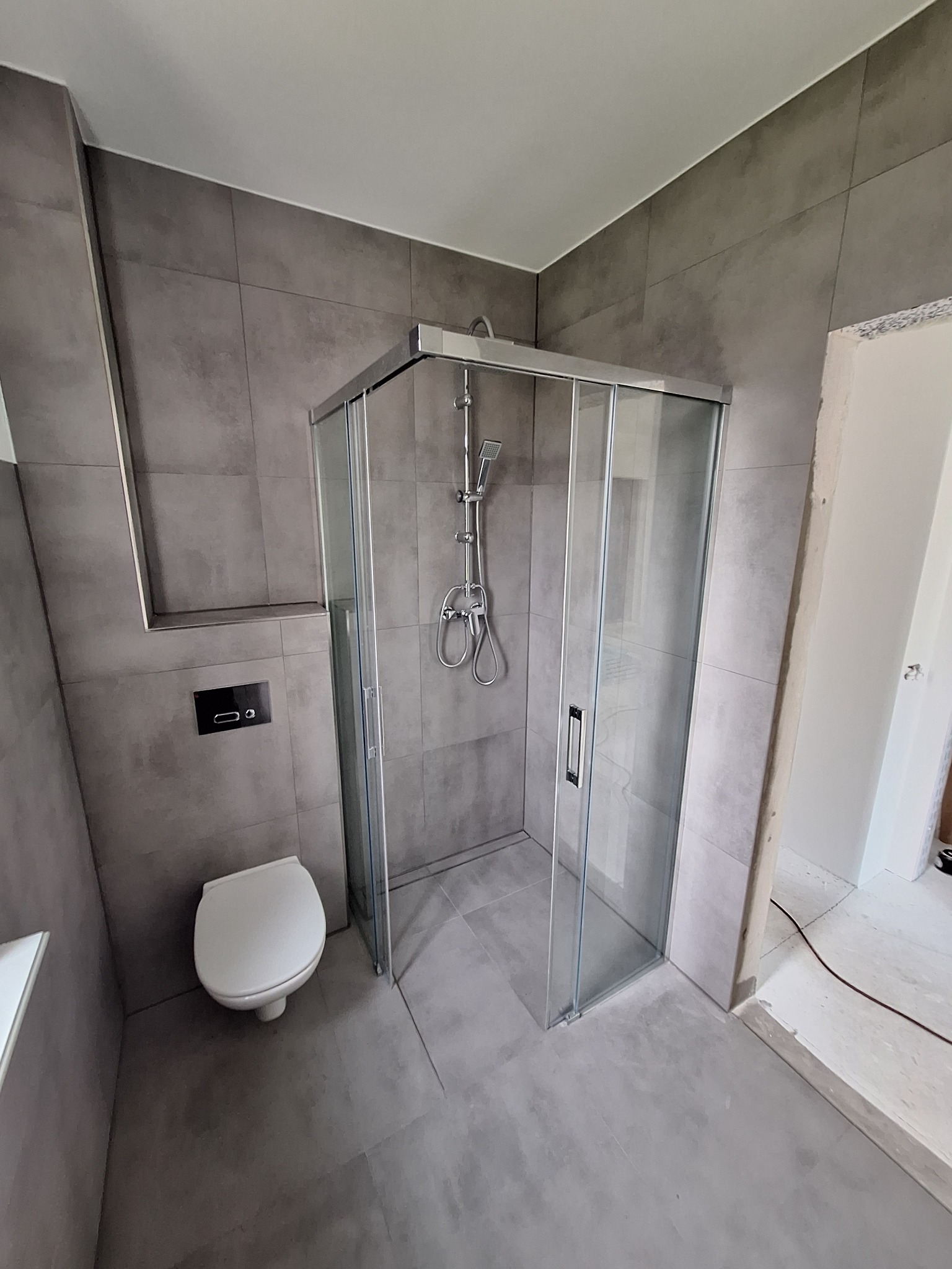 Nowoczesna łazienka z kabiną prysznicową z suwanymi drzwiami, wiszącą toaletą i szarą, matową glazurą w dużym formacie, widok na fragment niedokończonego przejścia do innego pomieszczenia.