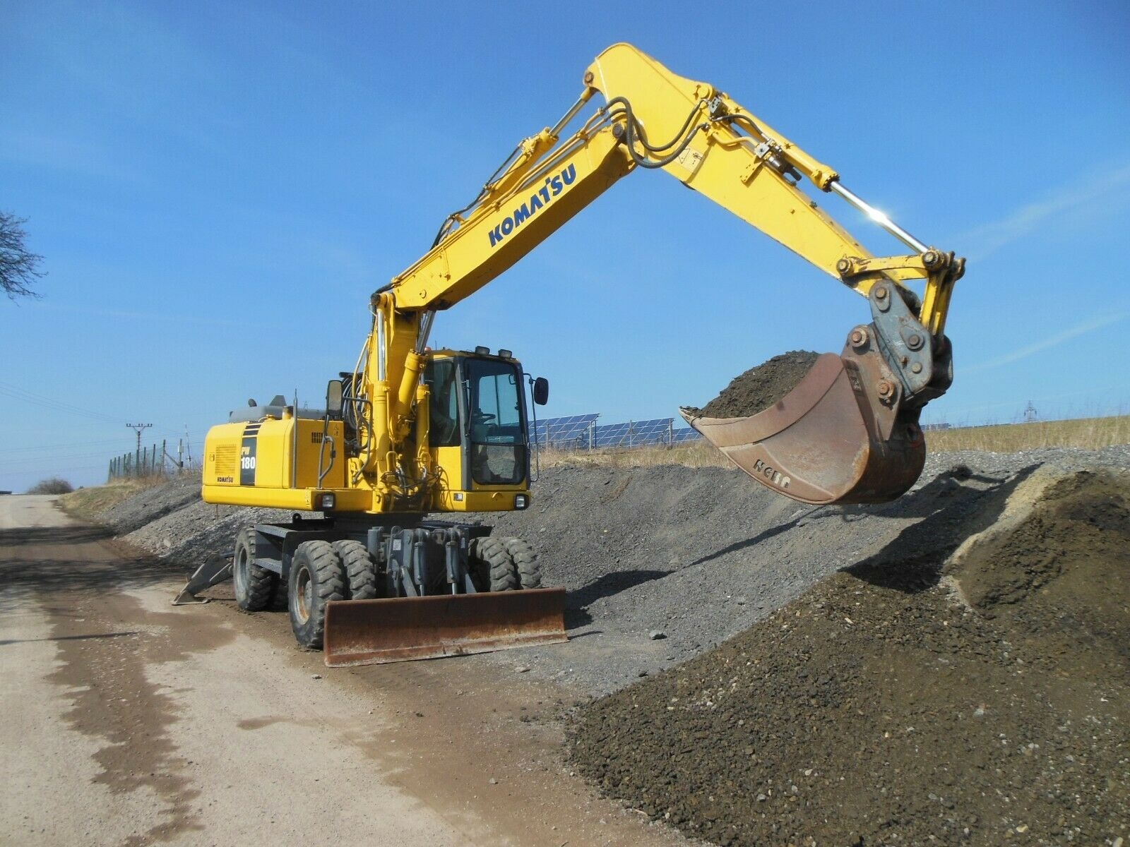 Żółta koparka kołowa Komatsu PW180 nabiera ziemię z pryzmy, w tle panele słoneczne i błękitne niebo.