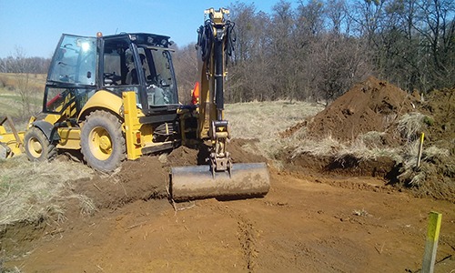 Żółta koparko-ładowarka podczas prac ziemnych na terenie z drzewami w tle, widoczny wykop i pryzma ziemi.