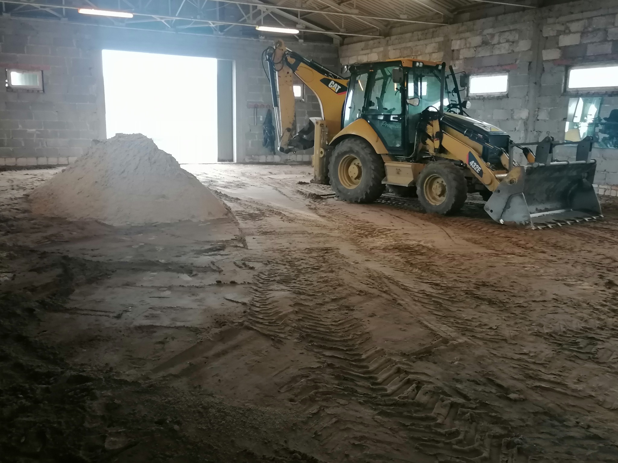 Żółta koparko-ładowarka Caterpillar 432E stoi wewnątrz budynku z betonowych bloków, obok pryzmy piasku, widoczne ślady opon na ziemi.